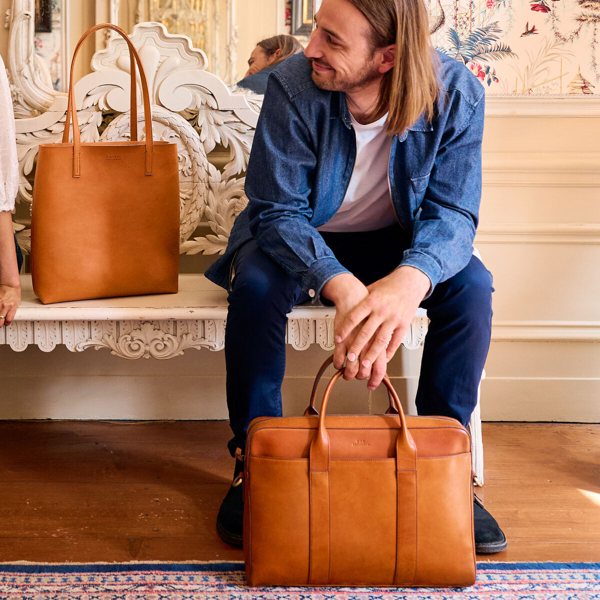 Ein Mann und eine Frau sitzen nebeneinander in einem elegant eingerichteten Raum. Beide halten Ledertaschen des Modells »Harvey«. Die Frau trägt eine weiße Bluse und blaue Schlagjeans, der Mann trägt eine blaue Jacke und Jeans. Die Szene strahlt eine gemütliche und modische Atmosphäre aus, unterstützt durch die luxuriösen Ledertaschen.