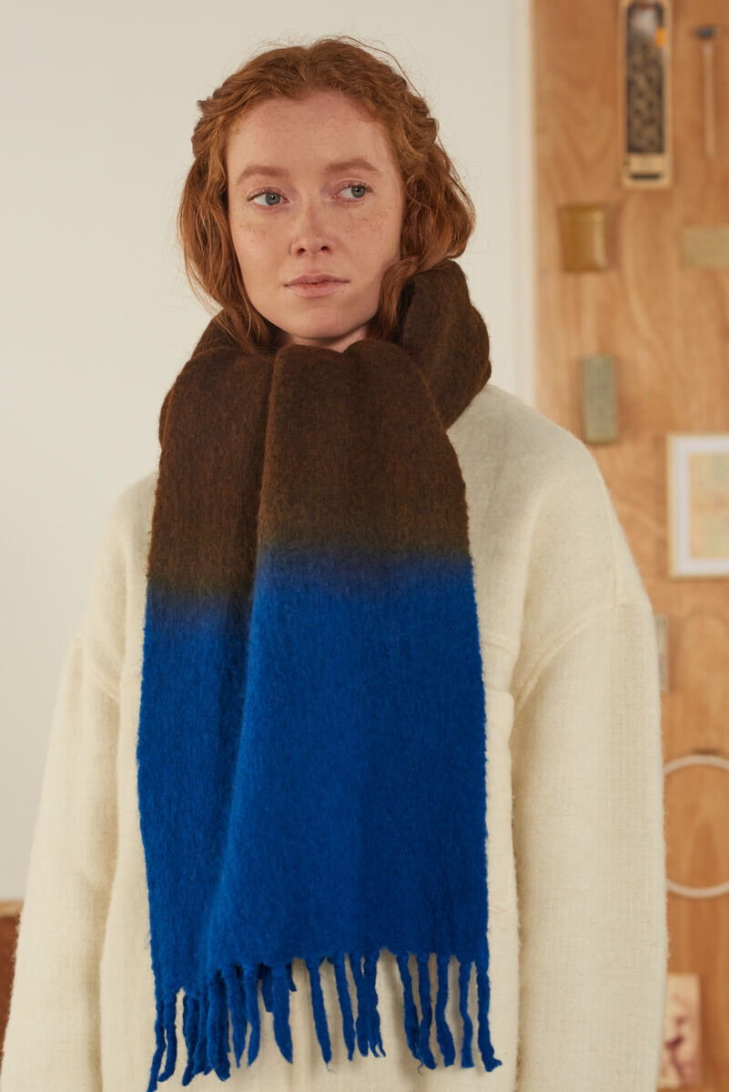 Eine Frau trägt einen dicken Schal, der oben braun und unten blau gefärbt ist. Der Schal ist handgestrickt und scheint sehr warm und kuschelig zu sein. Im Hintergrund sind Holzwände und Dekorationsgegenstände zu sehen.