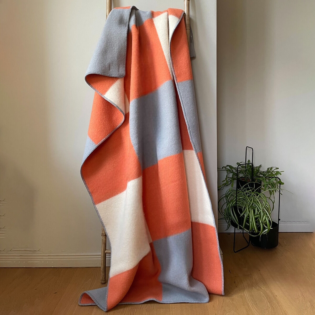 Eine dreifarbige Decke mit orange, grau und weißem Blockmuster hängt dekorativ über eine Holzleiter. Im Hintergrund steht eine grüne Pflanze. Sie bildet einen stilvollen und modernen Akzent für Wohn- oder Schlafzimmer.