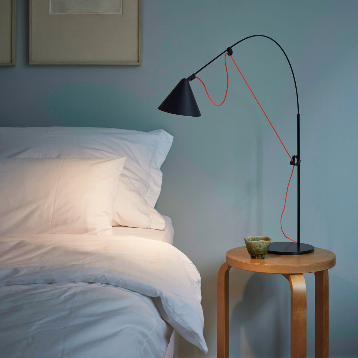 Ein modernes Schlafzimmer mit einer schwarzen Tischlampe »AYNO« auf einem runden Holztisch neben einem Bett mit weißer Bettwäsche. Die Lampe hat ein minimalistisches Design mit einem gebogenen Arm und einem orangefarbenen Kabel. Die Wand ist in einem hellgrauen Farbton gestrichen und es hängen zwei gerahmte Bilder darüber.