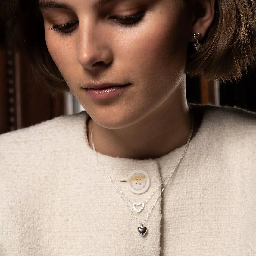 Nahaufnahme einer jungen Frau mit kurzer Frisur, die eine cremeweiße Jacke trägt und ein feines Collier mit zwei silbernen Herzanhängern um den Hals hat. Das Schmuckstück liegt dezent auf der Kleidung und wird elegant präsentiert.