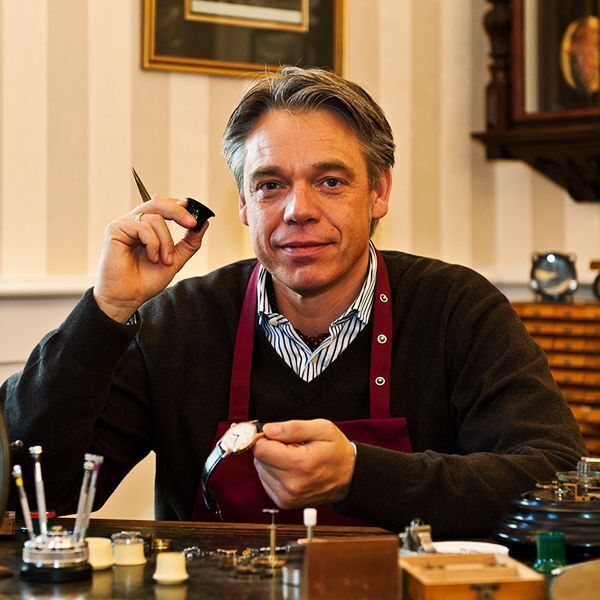 Ein Mann in einem dunkelbraunen Pullover und einer roten Schürze sitzt an einem Tisch mit Uhrmacherwerkzeugen und hält eine Edelstahl-Armbanduhr in der Hand.