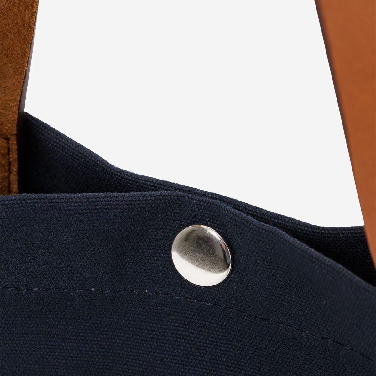 Nahaufnahme eines Teils einer Stoff-Einkaufstasche mit einem silbernen Knopf. Der obere Bereich der Tasche ist schwarz, und ein Teil des braunen Lederriemens ist sichtbar.