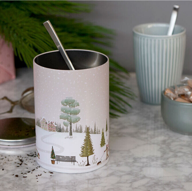 Ein Set von zwei zylindrischen Plätzchendosen mit einem weihnachtlichen Winterlandschaftsdesign. Eine Dose ist geöffnet und neben ihr eine graue Tasse mit Löffel, einem rosa Geschenk und einem kleinen Gefäß mit Karamellen. Die Szene ist auf einem grauen Marmortisch mit Tannenzweig im Hintergrund arrangiert.