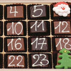 Nahaufnahme eines Adventskalenders von Gmeiner. Die einzelnen Fächer enthalten eckige Pralinen mit weißer Ziffernbeschriftung, dekoriert mit weihnachtlichen Motiven wie einem kleinen Santa-Gesicht und einem grünen Tannenbaum aus Zucker, eingelegt in einer eleganten Holzbox mit goldfarbenem Einsatz.