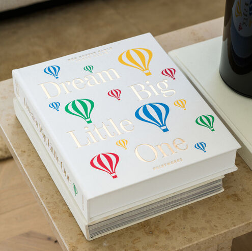 Auf einem Beistelltisch liegt ein Babyalbum mit dem Titel 'Dream Big Little One'. Das Cover des Albums ist weiß und mit bunten Heißluftballons in den Farben Rot, Gelb, Blau und Grün verziert.
