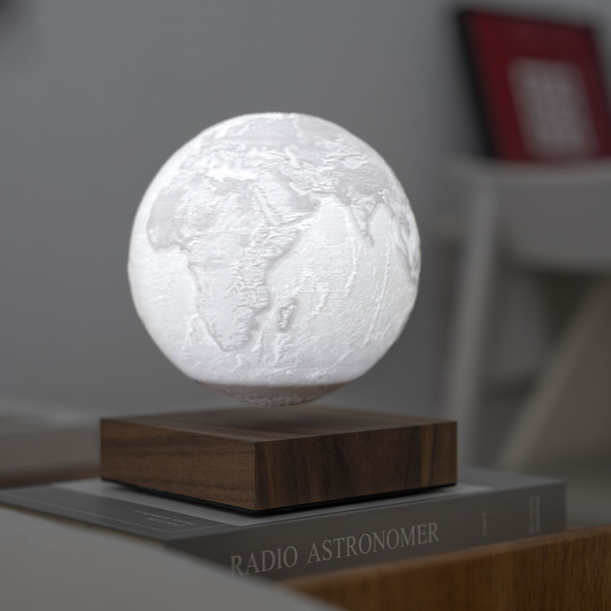 Die Gingko Smart Earth Lamp schwebt auf einem rechteckigen Holzsockel über einem Tisch. Die leuchtende Kugel zeigt eine detaillierte Weltkarte. Im Hintergrund sind unscharfe Bücher und Möbel zu sehen. Das Design ist modern, minimalistisch und technisch ansprechend.