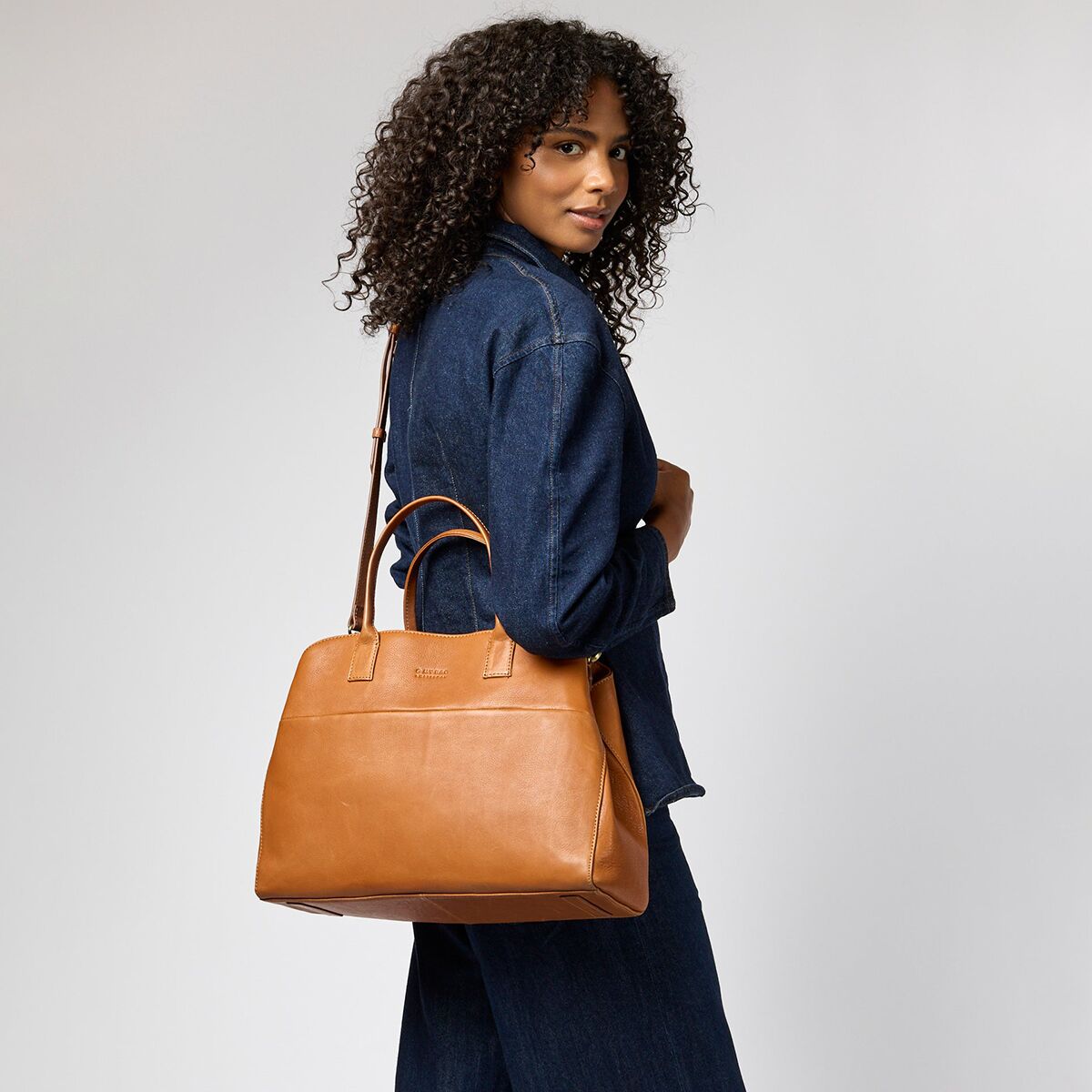 Eine Frau mit lockigem Haar trägt eine cognacbraune Schultertasche »Rita« von O My Bag aus glattem Narbenleder. Sie trägt einen dunkelblauen Jeans-Einteiler, steht seitlich und hält die stilvolle Tasche locker an ihrem Arm. Der Hintergrund ist neutral hellgrau.