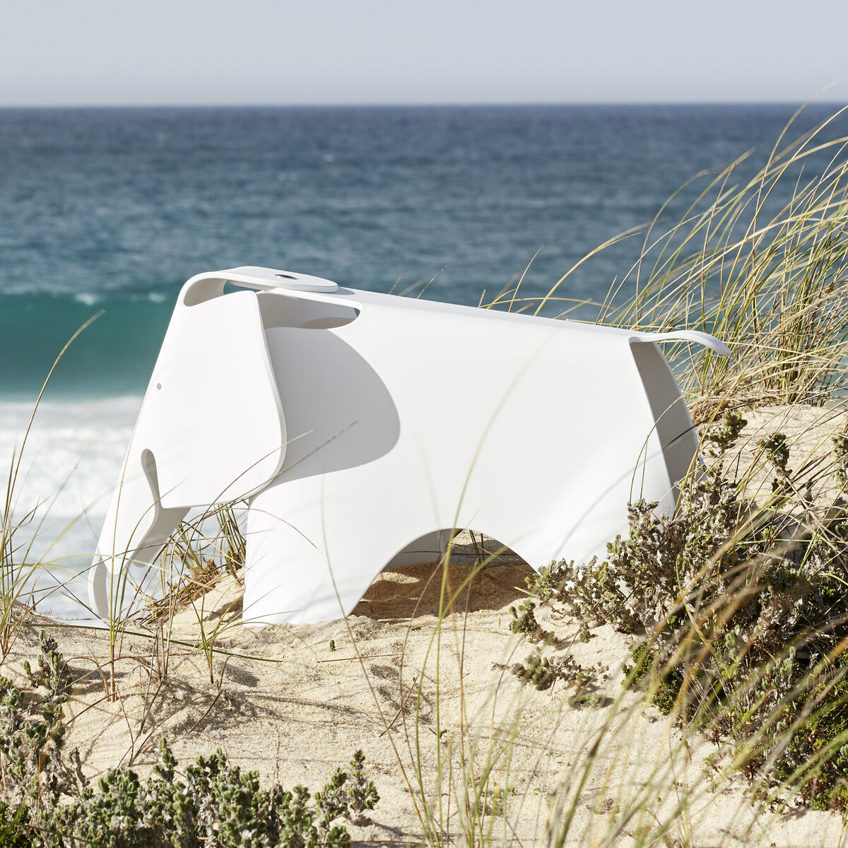 Ein weißer Eames Elephant Kinderhocker steht an einem Strand, umgeben von Dünen und Gras, mit Blick auf das blaue Meer.
