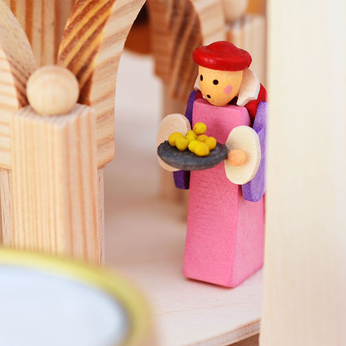 Eine detailreiche Weihnachts-Holzpyramide mit der Szene der Christgeburt. Eine der Figuren trägt Rosa und hält ein Tablett mit gelben Gegenständen, umgeben von Holzsäulen und weiteren kunstvollen Elementen. Die Pyramide ist für Teelichter und Pyramidenkerzen gestaltet.