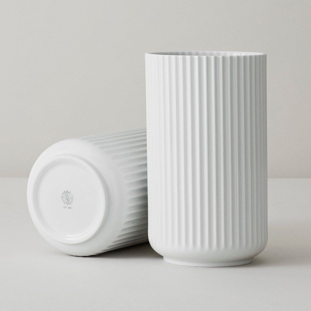 Zwei weiße, röhrenförmige Vasen »Lyngby« aus Keramik mit vertikalen Rillen. Eine Vase steht aufrecht, während die andere auf der Seite liegt, wodurch die Unterseite und das Logo sichtbar werden.