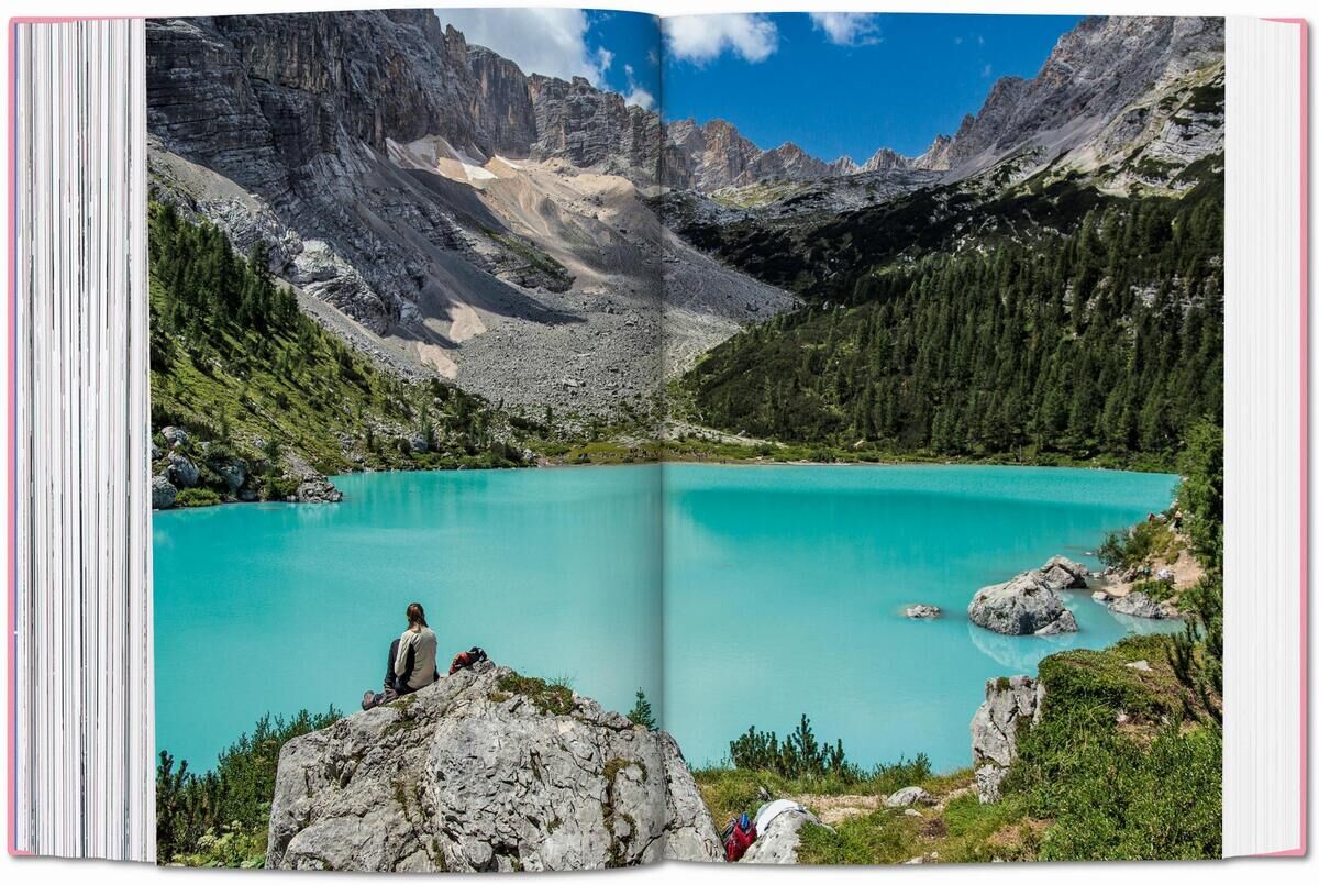 Ein aufgeschlagenes Buch mit dem Titel 'The New York Times Explorer. 100 Reisen rund um die Welt'. Die Doppelseite zeigt eine atemberaubende Landschaft mit einem türkisfarbenen See, umgeben von grünen Bäumen und grauen Berggipfeln. Eine Person sitzt auf einem Felsen am Ufer des Sees und genießt die Aussicht.