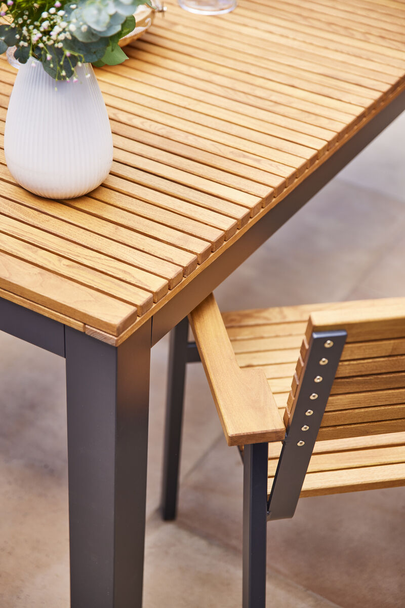 Ein moderner Holztisch mit Metallbeinen und passenden Holzstühlen steht auf einem leicht gemusterten Untergrund. Auf dem Tisch befindet sich eine weiße Vase mit grünen Pflanzen. Der Tisch hat eine rechteckige Holzoberfläche in warmem Braun.