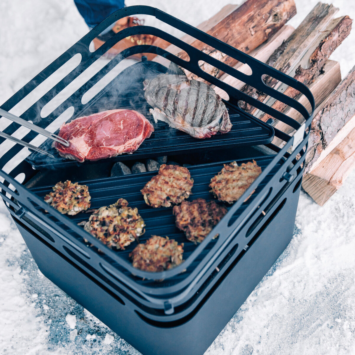 Ein schwarzer, quadratischer »CUBE« Plancha Grill im Freien, auf dem Fleischstücke und Gemüse gegrillt werden. Holz und rauchende Kohlen sind im Hintergrund zu sehen.