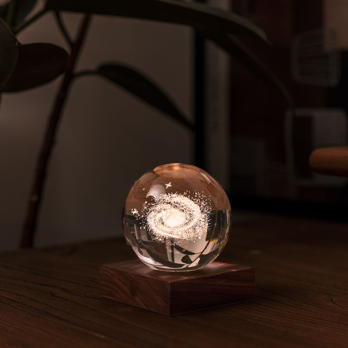 Eine dekorative Tischlampe in Form einer Kristallkugel mit weiigem Leuchteffekt und Holzsockel steht auf einer Holzoberfläche. Im Hintergrund sind unscharfe Pflanzen und Einrichtungselemente erkennbar. Die Lampe erzeugt ein warmes, gemütliches Ambiente.