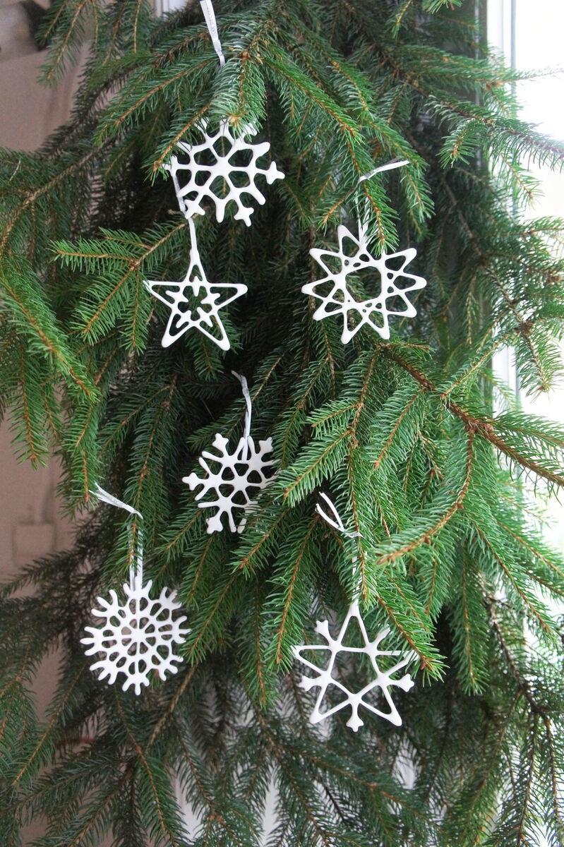 Sechs weiße, filigrane Porzellananhänger in Form von Sternen und Schneeflocken hängen an den Zweigen eines grünen Tannenbaums.