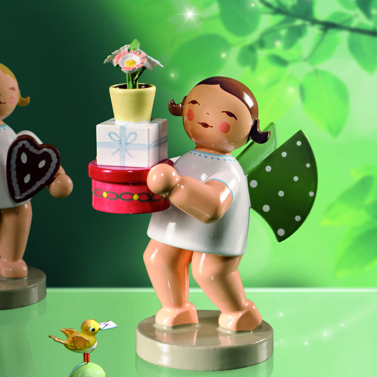 Zwei detailreiche Holzfiguren von Engeln der Marke Wendt & Kühn stehen auf glänzender Fläche vor grünem, von Lichtstrahlen durchzogenem Hintergrund. Ein Engel hält eine Torte, der andere einen Lebkuchen und Vogel. Am Boden ein gelber Vogel mit Brief.