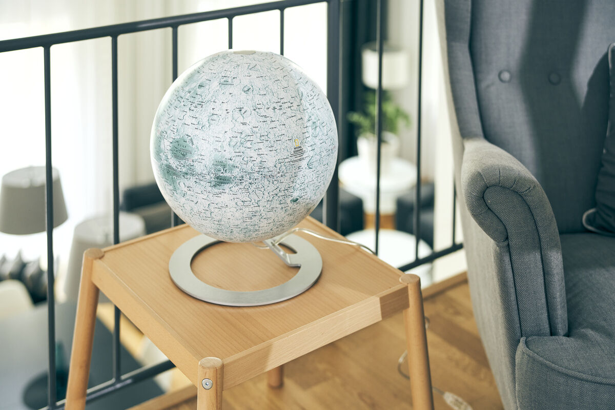 Ein Leuchtglobus von National Geographic „The Moon“ auf einem Holztisch im Wohnzimmer, der die Oberfläche des Mondes zeigt. Im Hintergrund sind ein Sofa und andere Einrichtungsgegenstände zu sehen.
