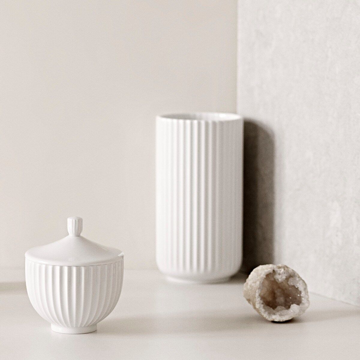 Zwei weiße Vasen im minimalistischen Stil stehen auf einem hellen, in beige gehaltenen Untergrund. Eine der Vasen hat einen Deckel, während die andere zylinderförmig ist. Im Hintergrund ist ein Kristall- oder Mineraliendekor zu sehen.