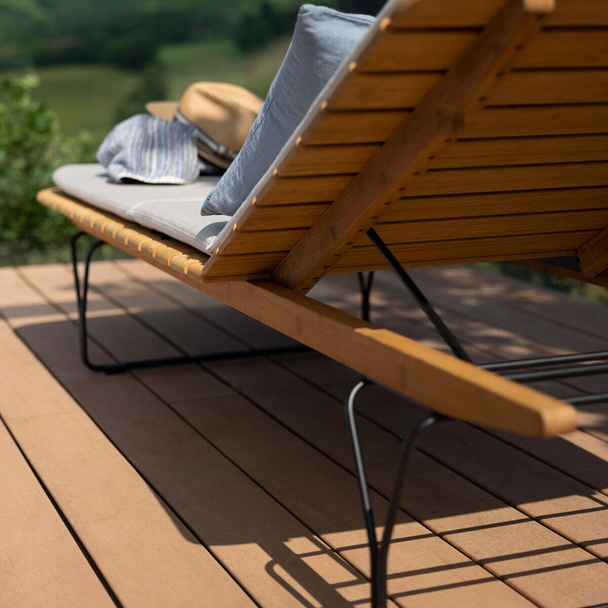Liege »MOLO« aus Holz auf einer Terrasse, mit grauen Kissen und einem Strohhut, im Hintergrund eine grüne Landschaft und blauer Himmel.