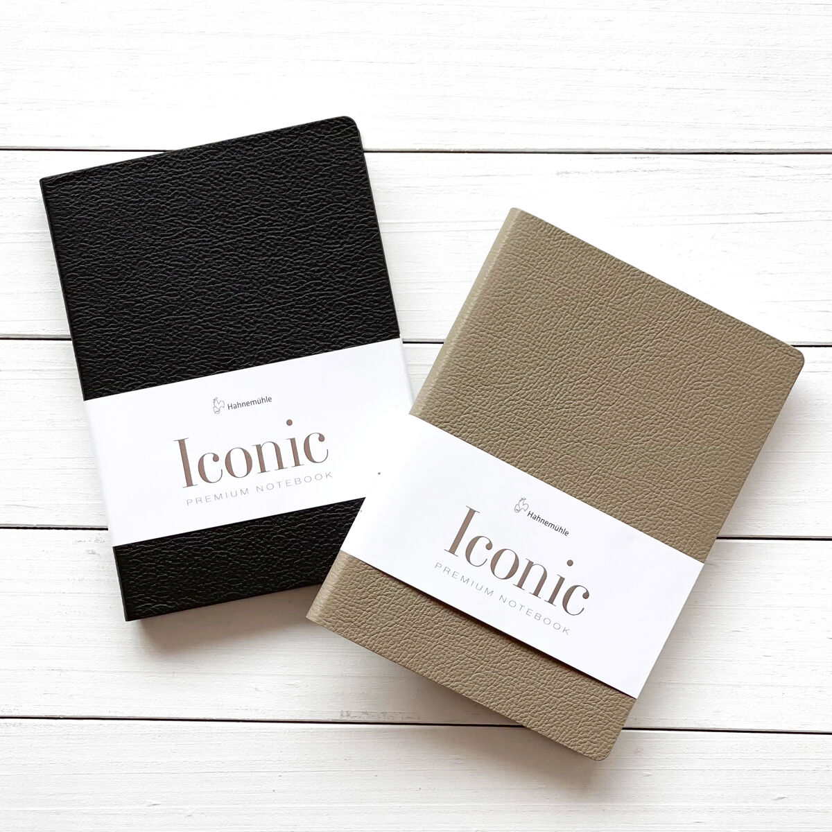 Zwei Notizbücher der Marke Iconic in DIN A5-Größe. Eines ist schwarz und das andere beige. Beide haben eine weiße Papierbanderole mit Aufschrift. Fotografiert auf einem weißen Holzuntergrund.