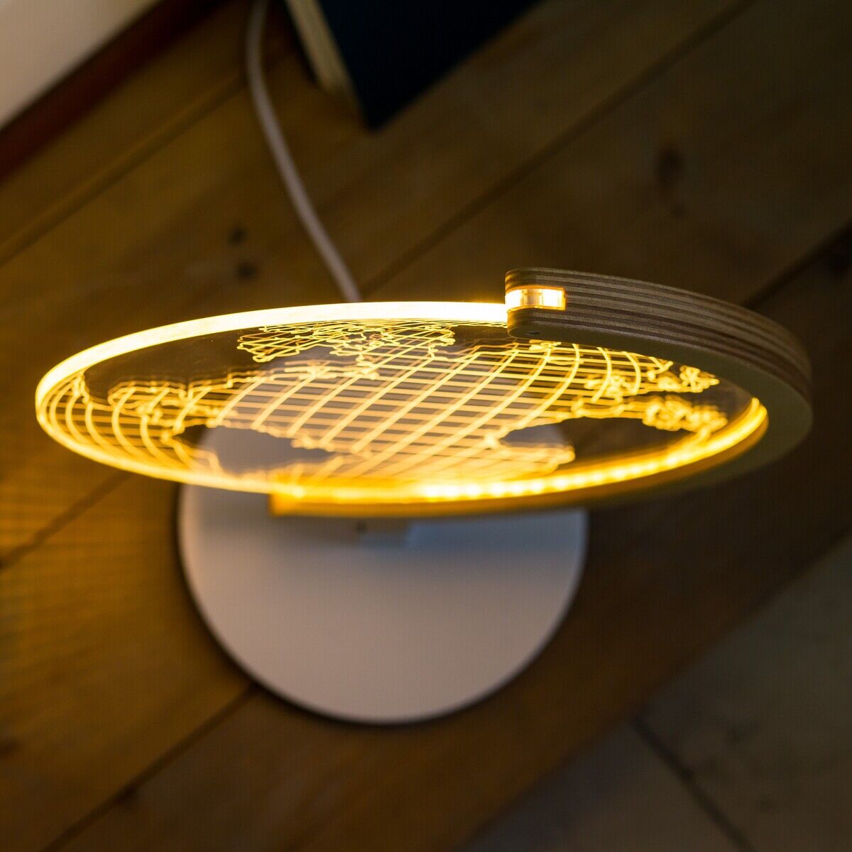 Eine LED-Leuchte in Form einer Weltkarte auf einem runden weißen Ständer, platziert auf einem Holzboden. Das Licht strahlt in warmem Gelb und erzeugt eine gemütliche Atmosphäre.