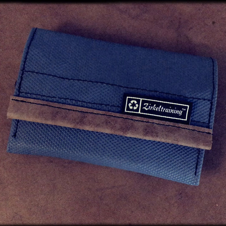 Bild einer blauen Geldbörse aus Leder mit dem Modellnamen Hechtrolle der Marke Zirkeltraining auf einer braunen Oberfläche. Die Geldbörse hat einen braunen Streifen und ein schwarzes Logo-Etikett.