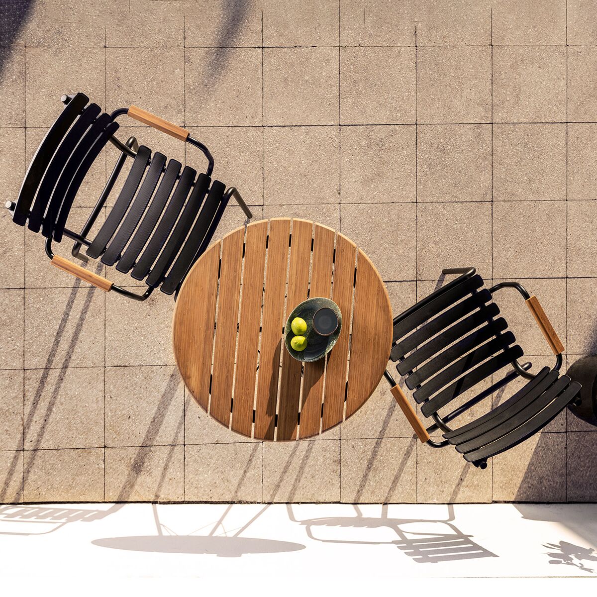 Draufsicht auf einen runden Holztisch mit Zitronen und einem Glas Wasser, umgeben von zwei modernen schwarzen Stühlen mit schwarzen Kunststofflamellen und Bambus-Armlehnen auf heller gefliester Terrasse. Sonnenlicht erzeugt markante Schatten am Boden.