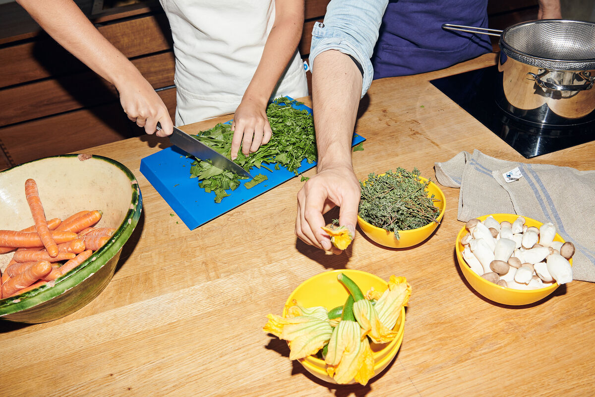 Zwei Personen schneiden grüne Kräuter auf einer blauem Schneidebrett mit einem Küchenmesser aus rostfreiem Stahl. Auf der Holzarbeitsplatte gibt es eine grüne Schüssel mit Karotten, gelbe Schüsseln mit Gemüse und Kräutern sowie einen Edelstahltopf.
