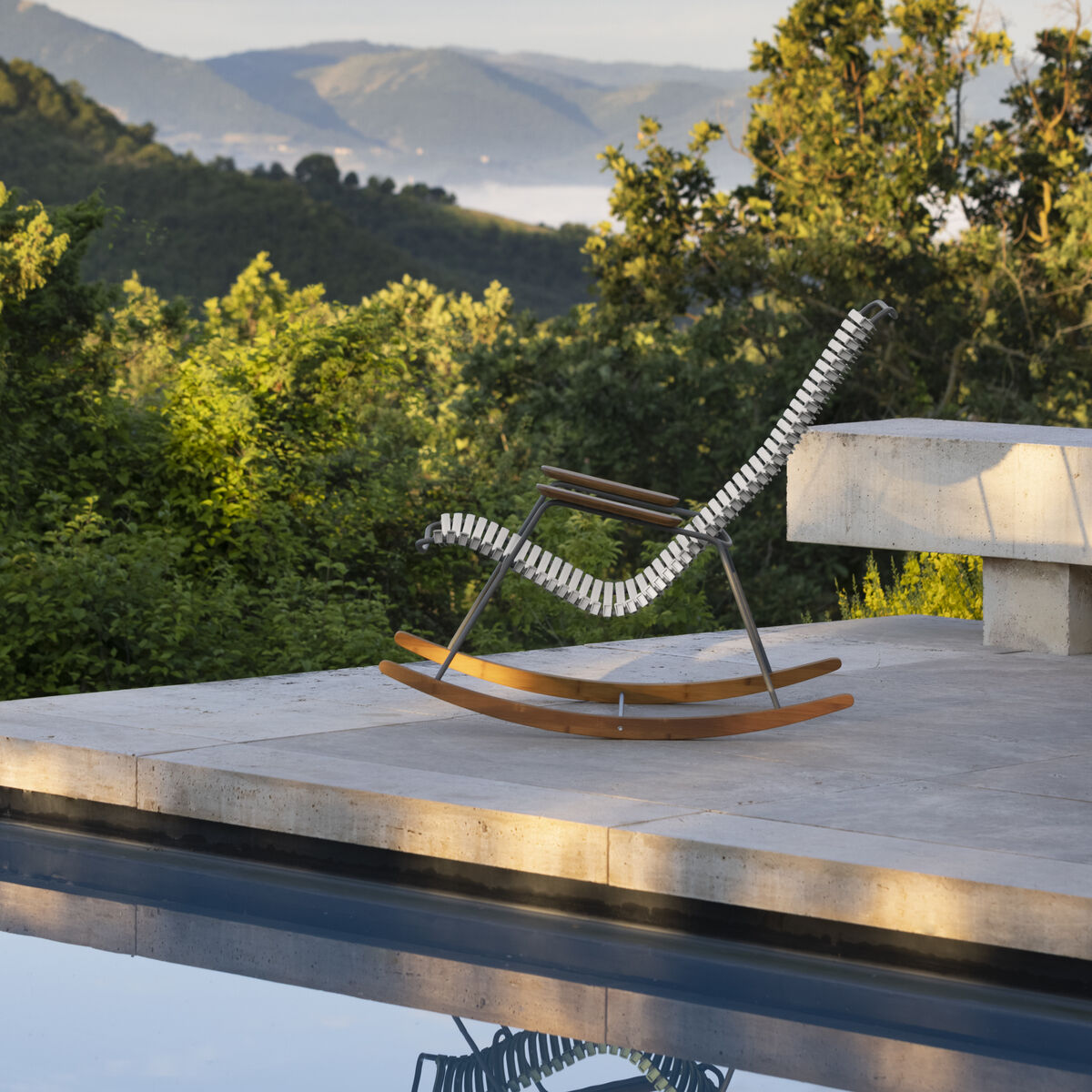 Schaukelstuhl »CLICK Rocking Chair« auf einer Terrasse mit Blick auf bewaldete Hügel und einen reflektierenden Pool im Vordergrund. Der Stuhl hat eine stilvolle, ergonomische Form mit natürlichen Holzelementen.