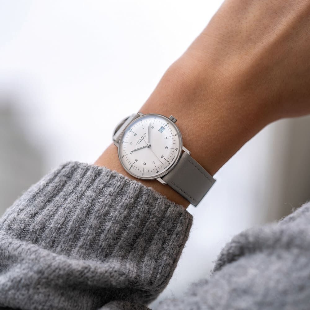 Eine minimalistische Armbanduhr aus der ZEIT-Kollektion »Max Bill Kleine Automatic« mit grauem Lederarmband, silbernem Edelstahlgehäuse und weißem Zifferblatt, getragen am Handgelenk einer Person mit grauem Strickpullover im Hintergrund.