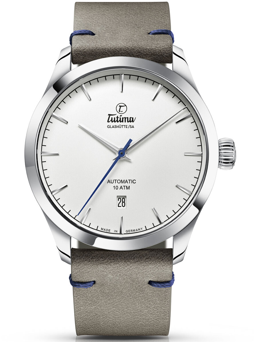 Dieses Bild zeigt die ZEIT Sonderedition »Flieger Aero Club« Automatikuhr von Tutima. Die Uhr hat ein Edelstahlgehäuse und ein graues Lederarmband mit blauen Nähten. Das Zifferblatt ist weiß mit silberfarbenen Markierungen und Zeigern.