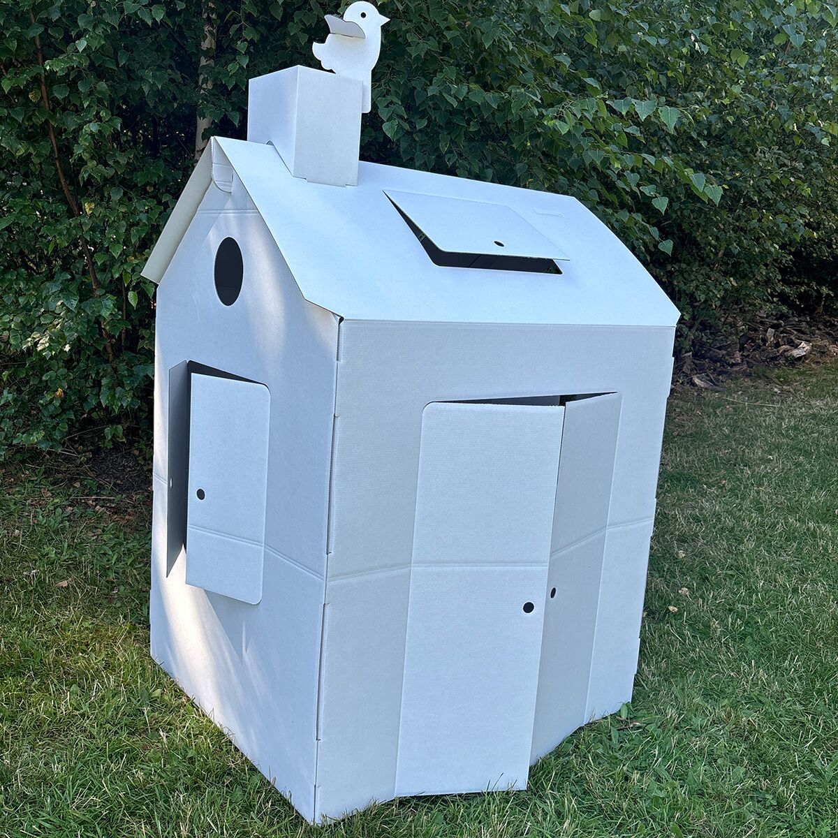 Ein großes, weißes Spielhaus aus stabiler Wellpappe steht auf einer Rasenfläche vor dichtem grünen Buschwerk. Das Haus hat eine Schornsteinfigur in Vogelform, eine Tür, Fenster und Klappen. Es ist noch unbemalt und besteht aus mehreren Pappteilen, die ineinander gesteckt sind.