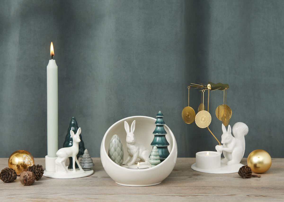 Ein Weihnachtsarrangement, das eine brennende Kerze, ein Eichhörnchen mit einem Engelsglockenspiel, einen Hirsch und einen Hasen unter Tannenbäumen in weißen Porzellanskulpturen zeigt. Das Arrangement enthält weihnachtliche Farben wie Weiß, Gold und Grün und ist auf einer Holztischoberfläche vor einem grauen Hintergrund platziert.