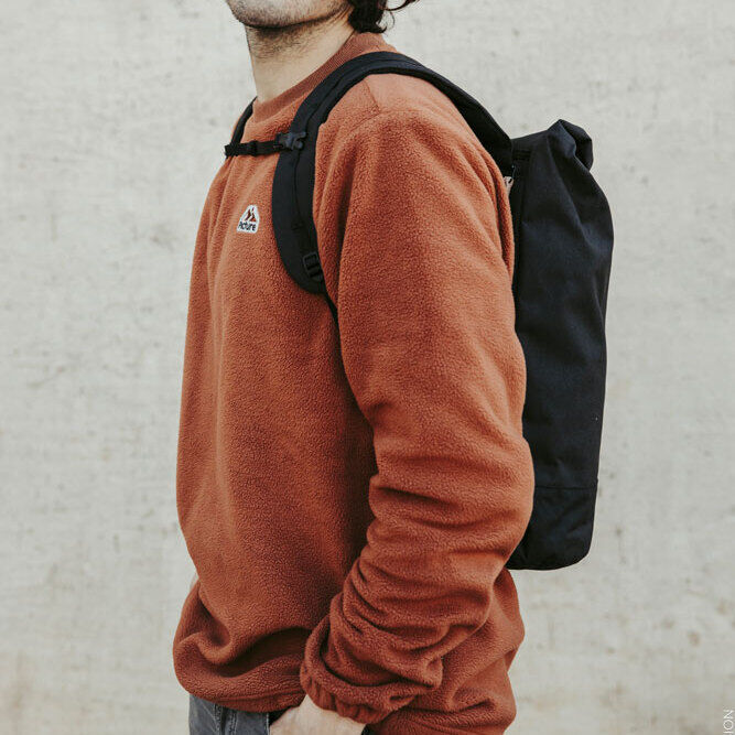 Ein Mann trägt einen schwarzen Rucksack/Fahrradtasche »Annecy« auf dem Rücken. Er hat eine Sonnenbrille und ein Stirnband an. Er trägt einen rostfarbenen Pullover und graue Jeans. Der Hintergrund ist eine helle, beige Wand.