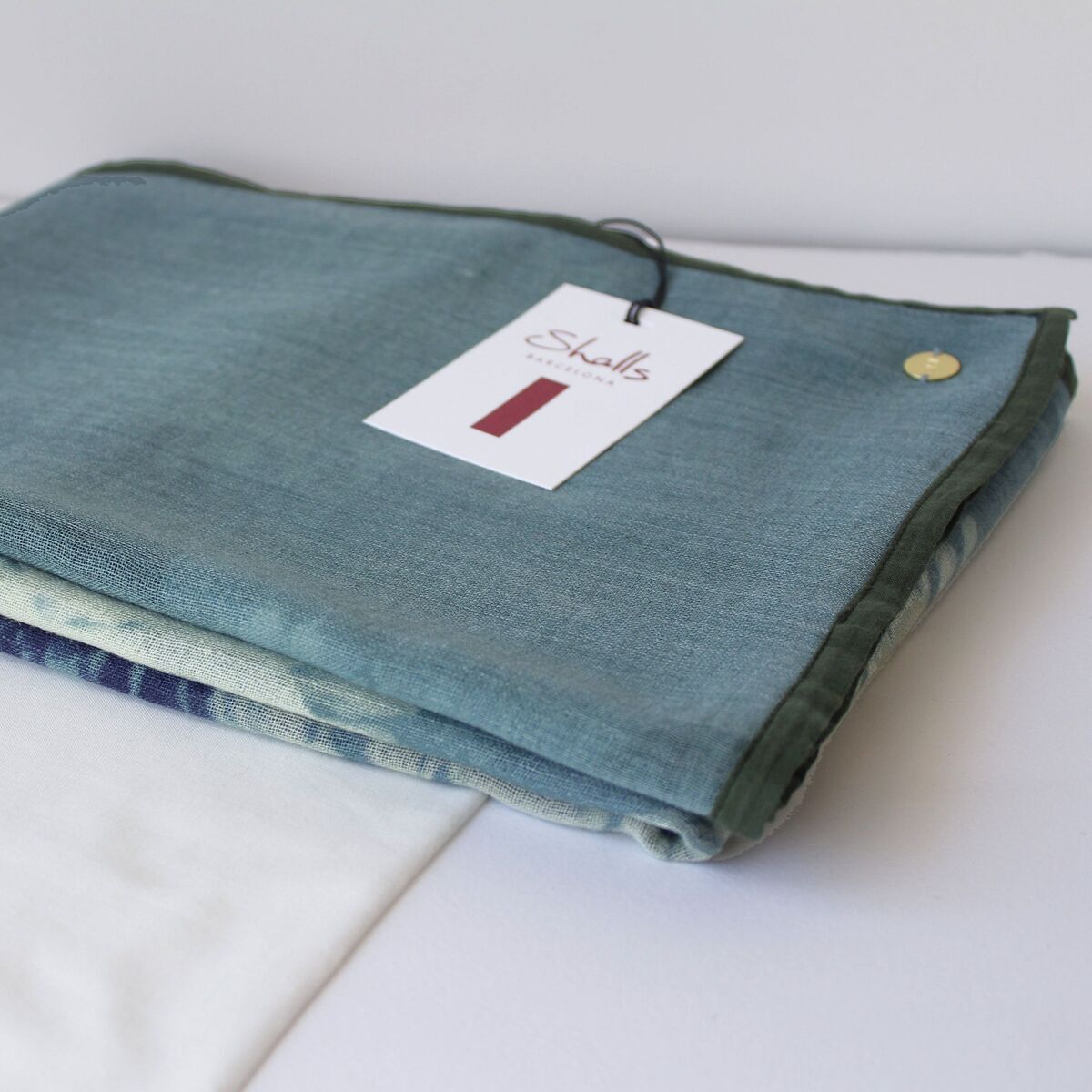 Ein eleganter Wollschal in eisblau liegt auf einer weißen Stofftasche. Der Schal wirkt weich und hochwertig verarbeitet. Daneben befindet sich ein Etikett mit Produktinformationen. Das Bild betont die feinen Materialdetails und das schlichte, zeitlose Design.