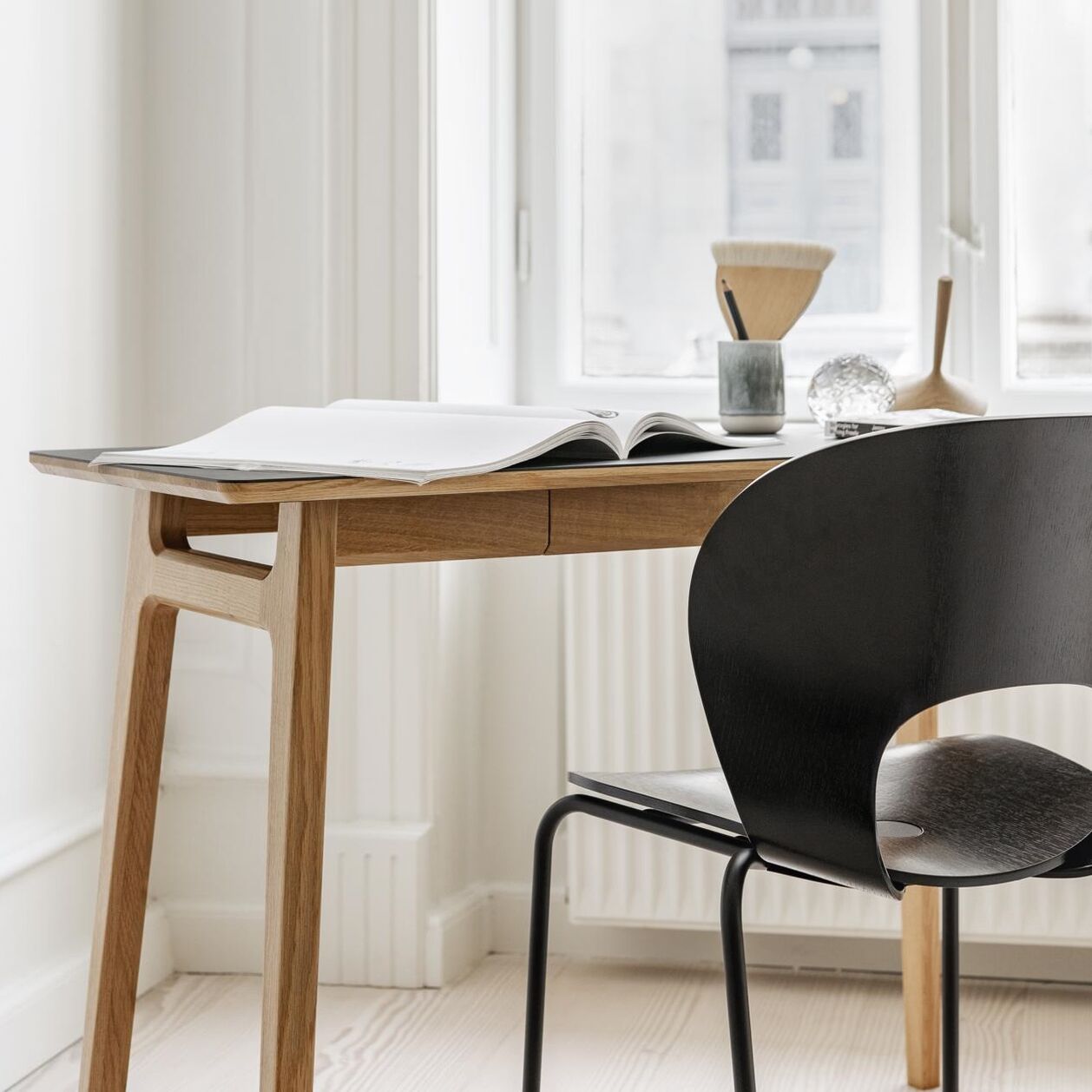 Eleganter Dining Chair »Ø« von Magnus Olesen in tiefem Schwarz, mit gebogener Rückenlehne aus Eiche, steht an einem hellen Holzschreibtisch mit aufgeschlagenem Buch und Stiftbechern. Heller Raum mit Holzfußboden und großem Fenster im Hintergrund.