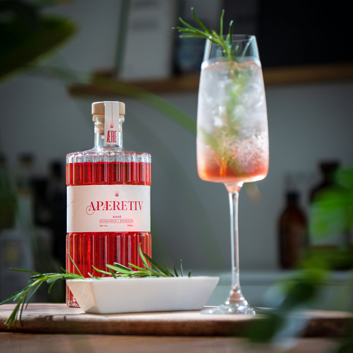 Eine Flasche AERE Aperitif »Rosé« steht auf einem Tisch neben einem gefüllten Weinglas, das mit Eis, Rosmarinzweigen und dem Getränk dekoriert ist.