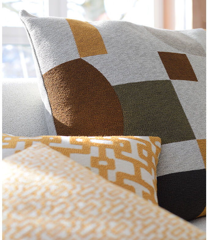 Ein Kissen mit geometrischem Muster in Weiß, Braun, Ocker und Dunkelgrau liegt auf einem Sofa. Neben ihm befindet sich ein weiteres Kissen mit gelbem und weißem Design.