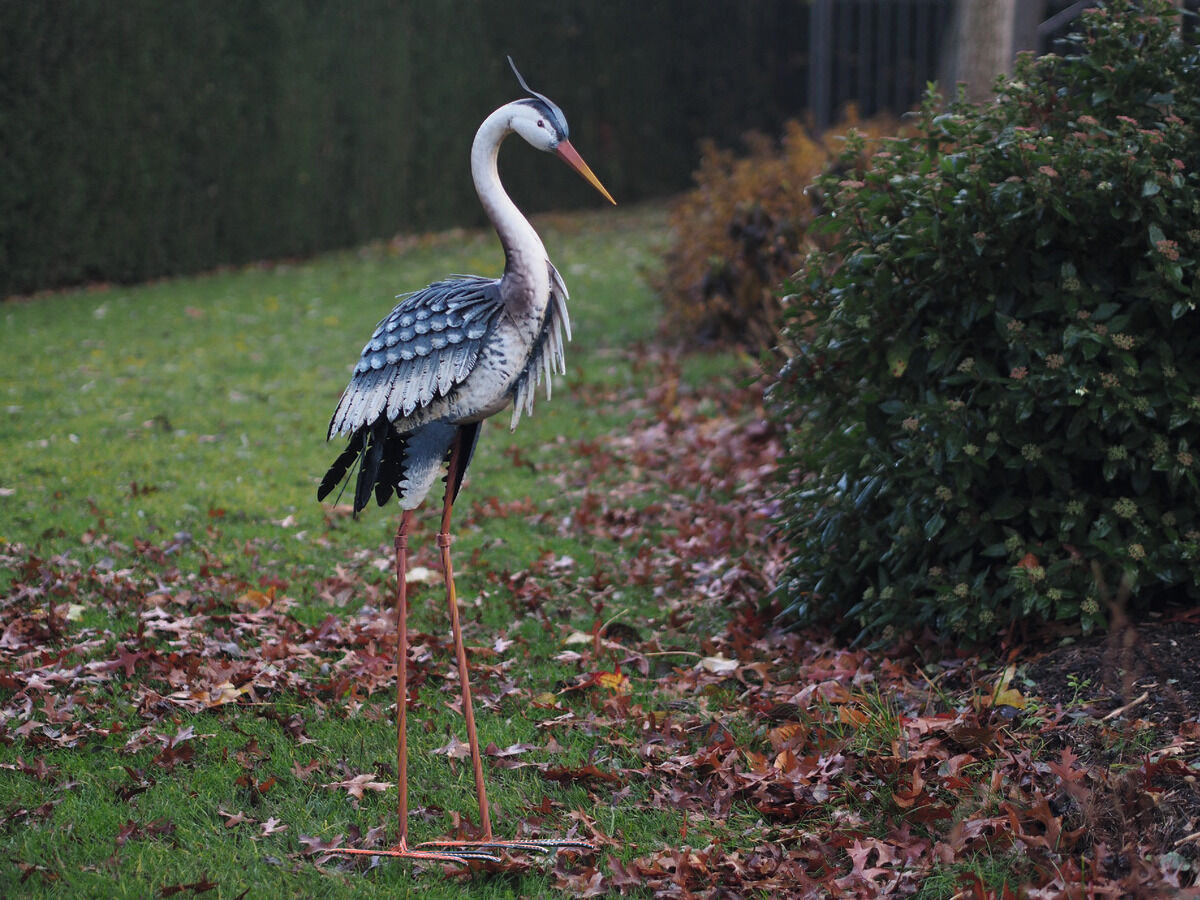 Eine realistisch gestaltete Gartenfigur eines Fischreihers steht auf einem Rasen, der von heruntergefallenem Herbstlaub bedeckt ist. Die Figur zeigt feine Details und ist überwiegend in Grün-, Braun-, Weiß- und Schwarztönen gehalten.