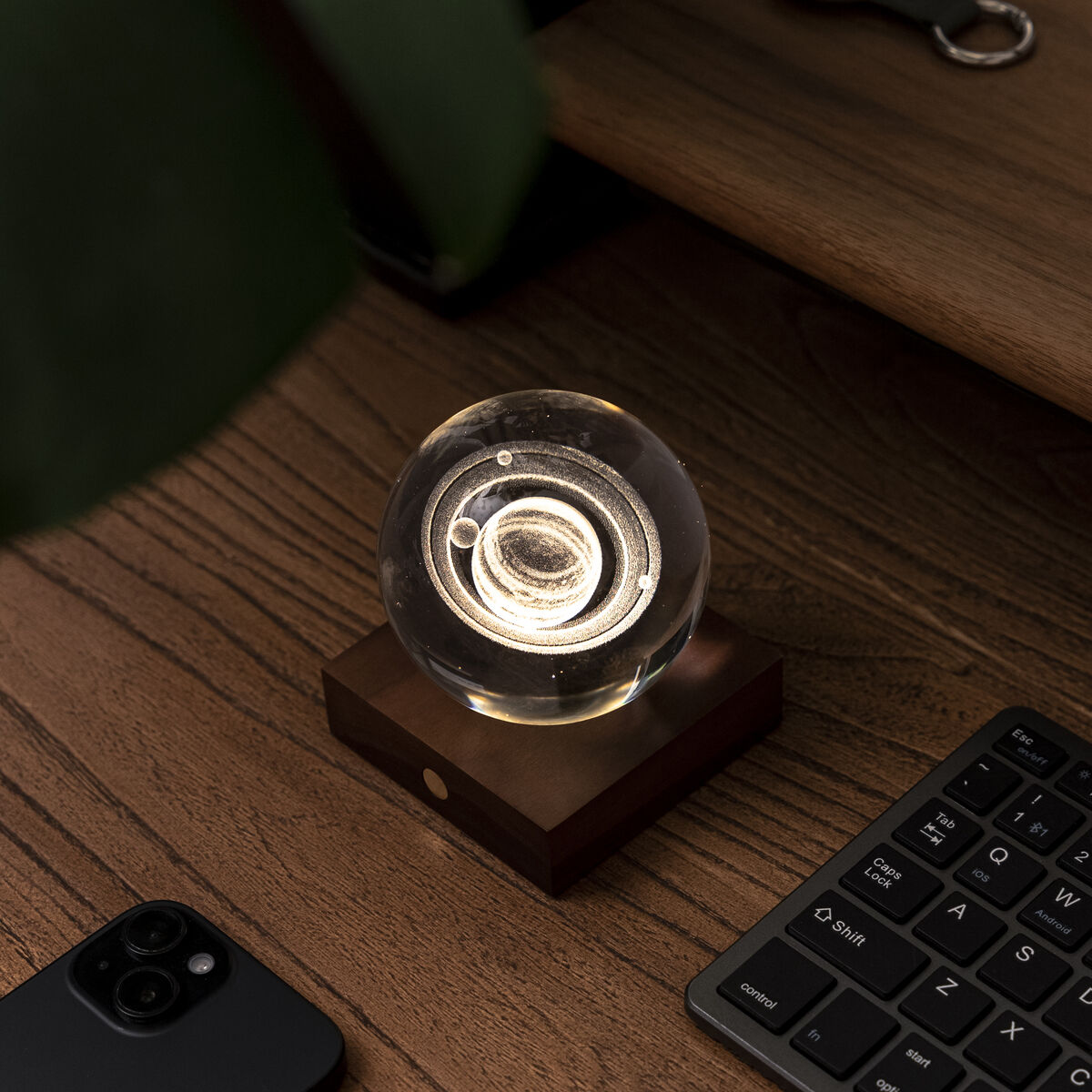 Eine moderne Glaskugellampe mit warmem Licht steht auf einem hölzernen Sockel auf einem Schreibtisch. Umgeben sind Tastatur, Smartphone und Pflanzen, was eine gemütliche, elegante und minimalistische Atmosphäre vermittelt.