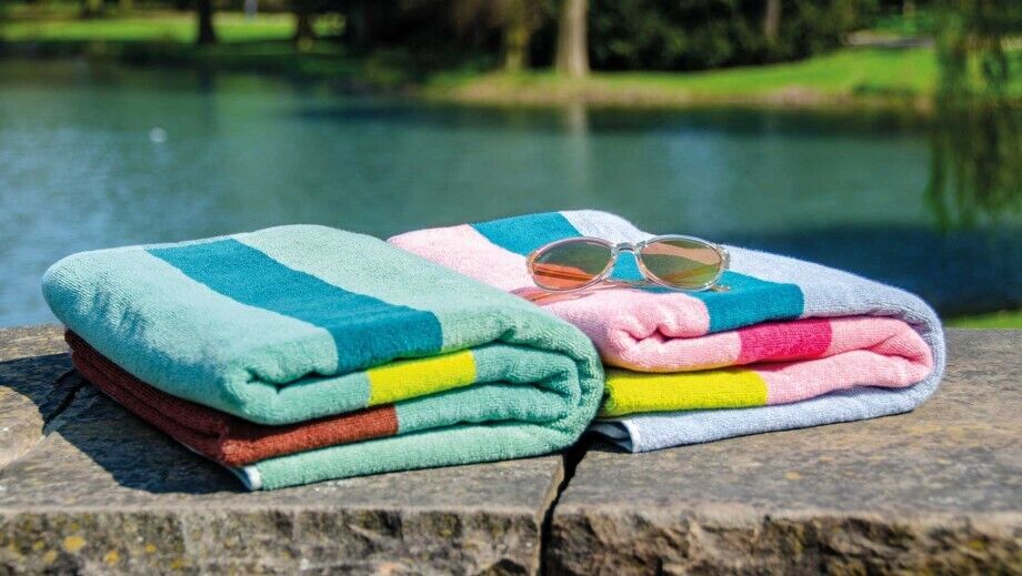 Zwei gefaltete Badetücher mit farbigen Streifen in Grün, Rosa, Blau und Gelb liegen auf einem Stein am Wasser, zusammen mit einer Sonnenbrille.