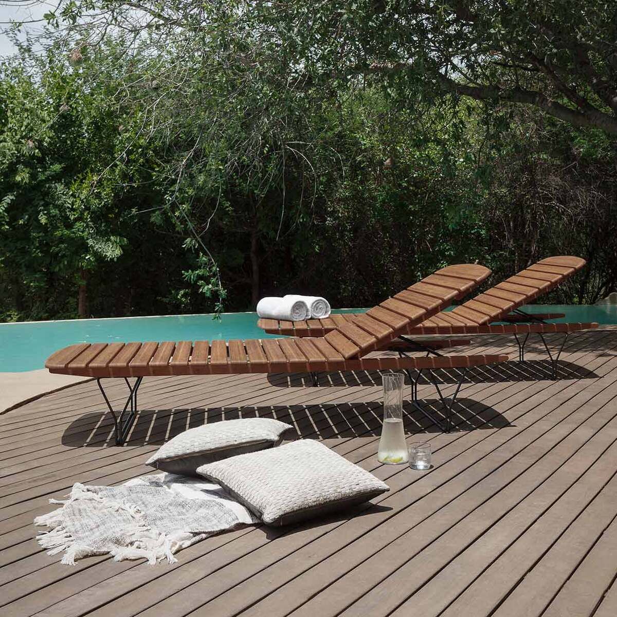 Zwei braune Holzliegen stehen auf einer Terrasse neben einem Pool. Im Vordergrund liegen zwei Kissen und ein Getränk mit einem Handtuch darauf. Bäume und grüne Sträucher umgeben den Pool.