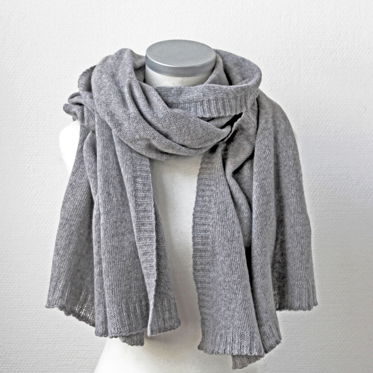 Eine graumelierte Kaschmirstola namens "Donna" ist locker und voluminös um eine weiße Schneiderbüste drapiert. Der Strickstoff wirkt weich und luxuriös. Der Hintergrund ist schlicht und weiß, wodurch das edle Material und die graue Farbe besonders hervorgehoben werden.