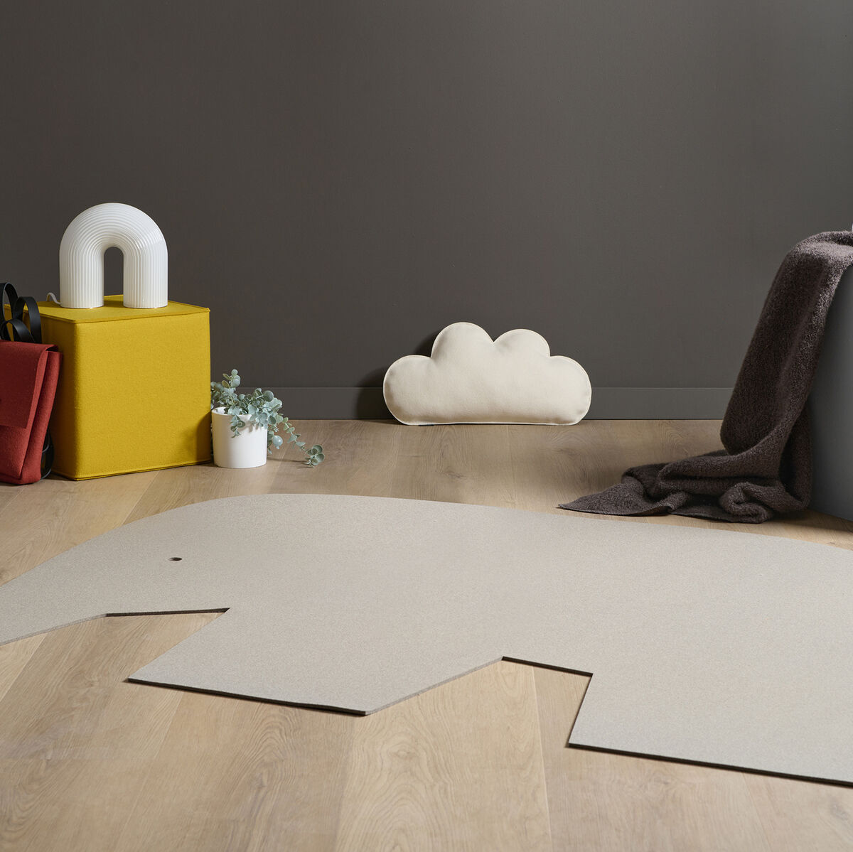 Ein stilvolles Kinderzimmer mit grauer Wand und Holzboden. Ein Wolkenkissen liegt auf dem Boden neben einem beigen Elefanten-Teppich. Auf einem gelben Würfel steht eine weiße Lampe, und ein kleiner Pflanzentopf ist daneben platziert. Ein grauer Stoff ist an einem anderen Würfel aufgehängt.