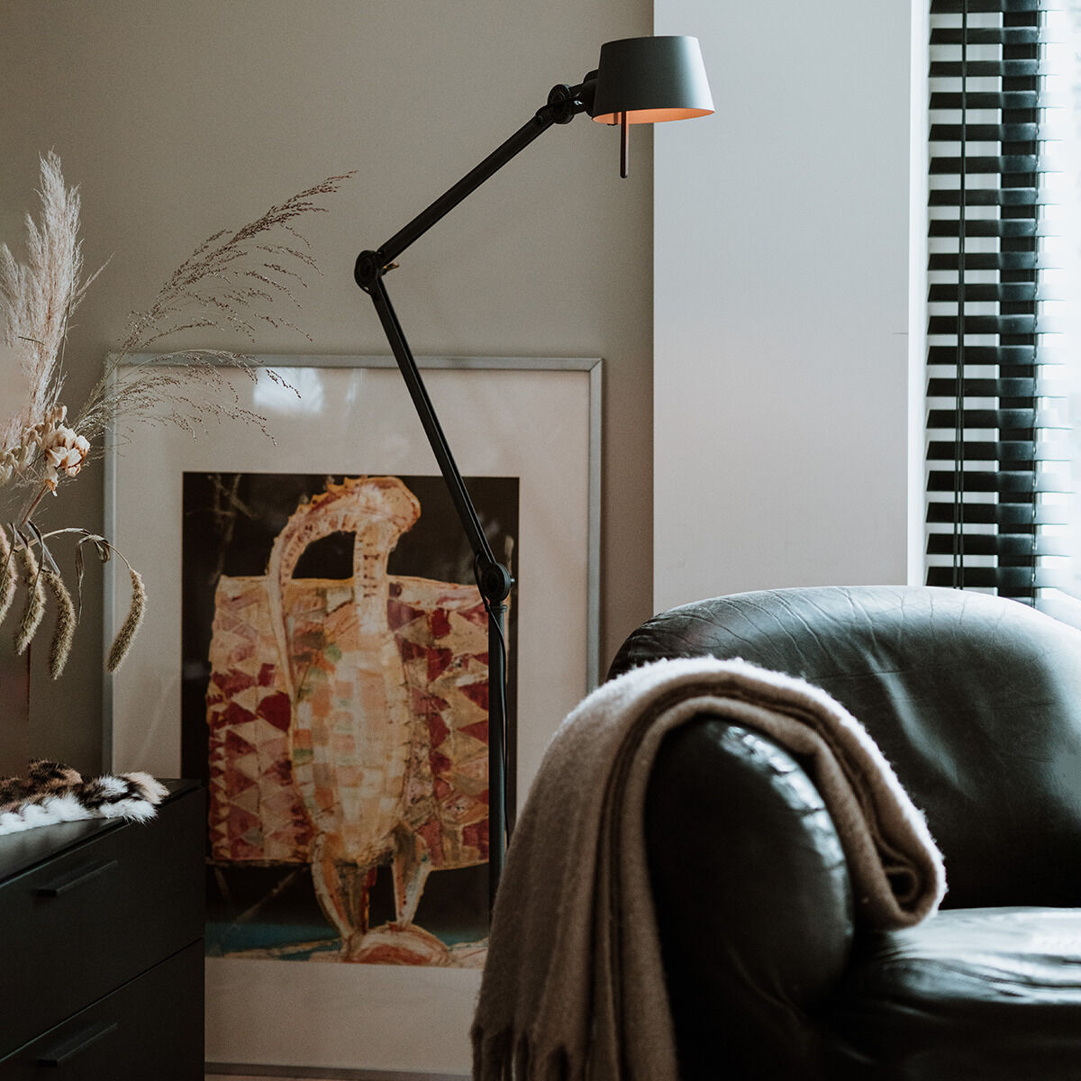 Eine moderne Stehlampe mit zwei schwenkbaren Armen steht in einer stilvollen Wohnzimmer-Ecke. Im Hintergrund ist ein künstlerisches Bild an der Wand zu sehen und neben dem schwarzen Beistelltisch befindet sich ein Ledersessel mit einer beigefarbenen Decke.