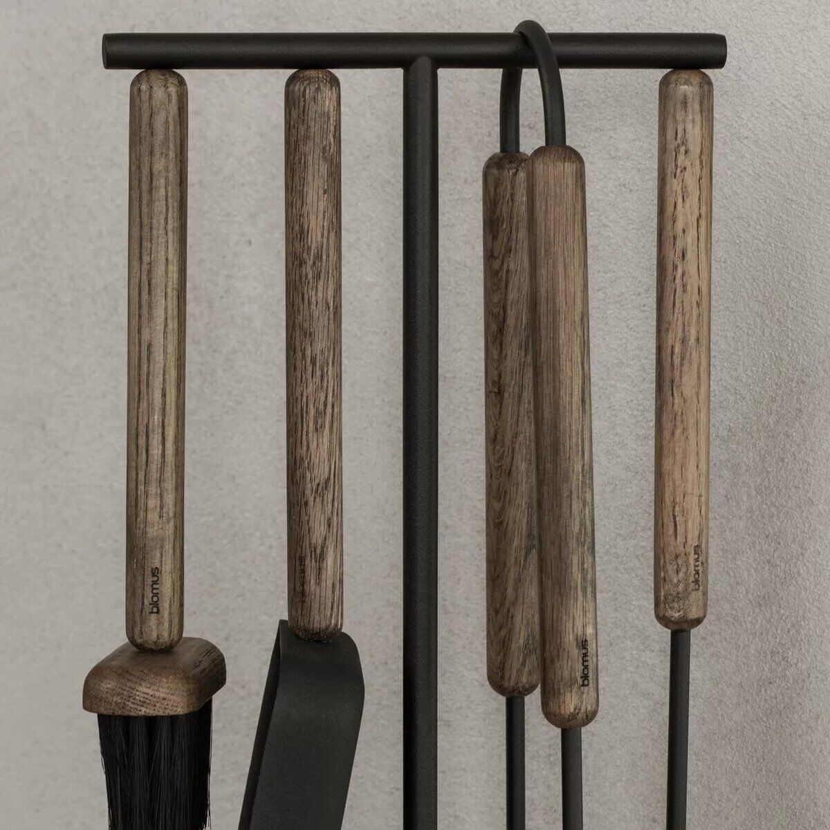 Modernes Kaminbesteck-Set mit Holzgriffen und schwarzen Metallstielen, bestehend aus Besen, Schaufel, Schürhaken und Zange. Die Werkzeuge hängen ordentlich an einem verticalen Metallständer vor einer hellgrauen Wand. Ideal für das Aufräumen und Pflegen eines Kamins im Wohnzimmer.