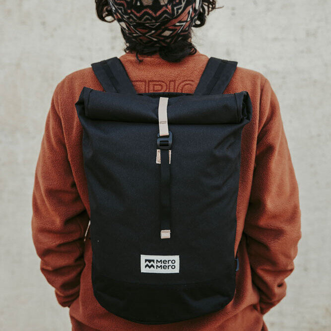 Person trägt eine schwarze Fahrradtasche/Rucksack »Annecy« auf dem Rücken, mit einer orangefarbenen Jacke und einer gemusterten Mütze.