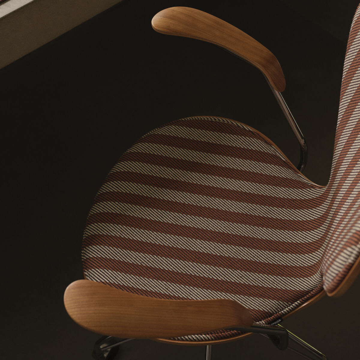 Detailansicht des Schreibtischstuhls 'Serie 7' von Fritz Hansen mit gestreiftem Bezug in Braun- und Beigetönen, hölzernen Armlehnen und Metallgestell. Der Stuhl steht vor einem Schreibtisch mit Papierstapeln unter warmem Licht in einer stilvollen Umgebung.