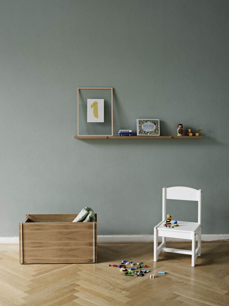 Ein Eichenholz-Wandregal an einer grauen Wand, dekoriert mit einem gerahmten Bild, einem Spielzeugbus und einem kleinen Bilderrahmen. Neben dem Regal steht eine weiße Kindersitzbank und eine Holzspielzeugkiste. Spielzeugteile sind auf dem Hartholzboden verstreut.